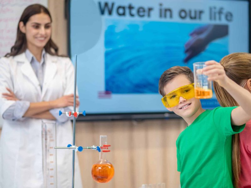 Entertaining lesson. Boy in goggles showing glass of colored liquid standing with back to girl and smiling woman in lab coat